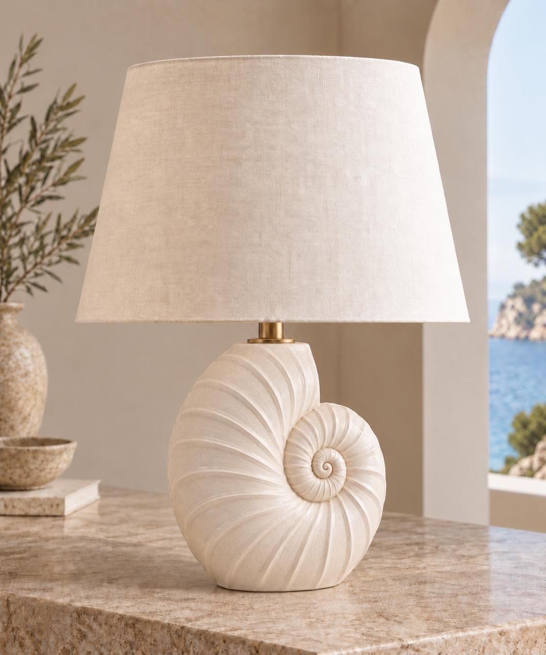 Lámpara de cerámica blanca en forma de caracol nautilo, sobre una mesa de mármol frente a una ventana al mar